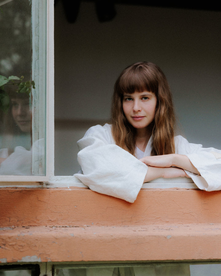 A woman rests her arms on an orange windowsill, framed by plants and soft natural light, adding a peaceful, intimate feel.