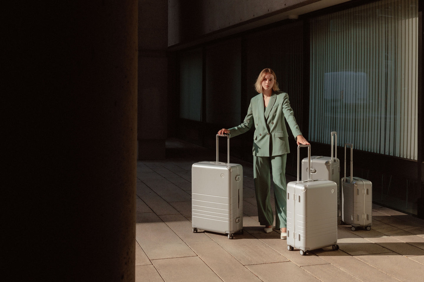 A woman in a green suit stands outdoors with four hybrid suitcases in silver of varying sizes, all with extended telescopic handles.