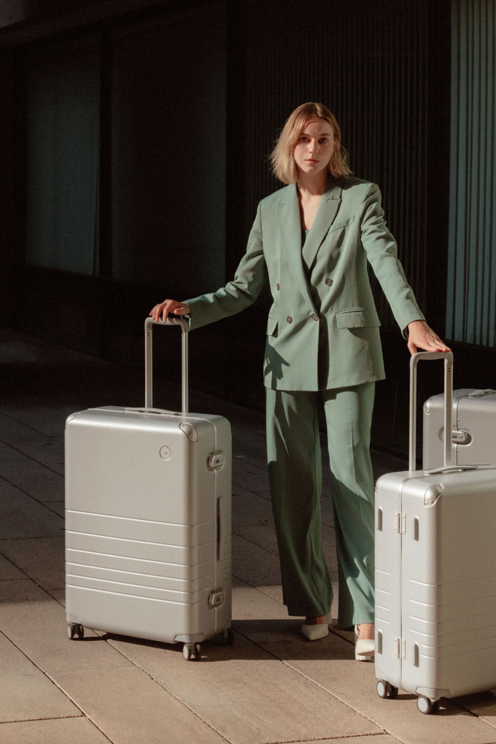 A woman in a green suit stands outdoors with three hybrid suitcases in silver of varying sizes, all with extended telescopic handles.