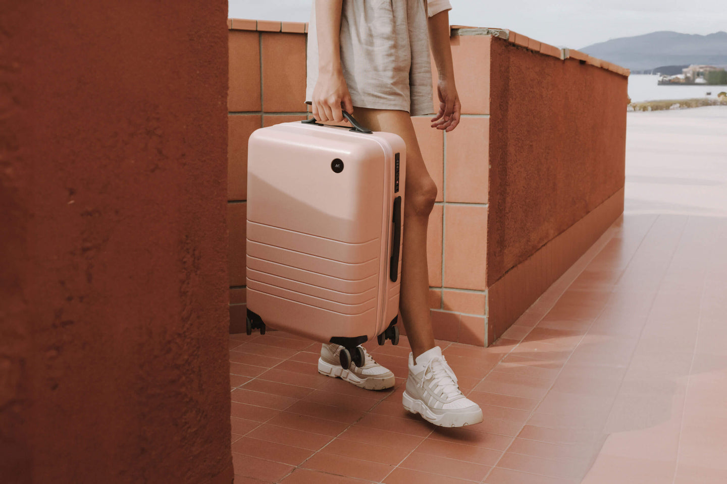 A person in casual summer attire walks along a terracotta-tiled corridor, carrying a Carry-On in Rose Quartz.