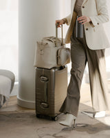 A woman dressed in a cream-coloured blazer and grey pants rolling a Hybrid carry-on in champagne, with a matching metro duffel in ivory (vegan leather) stacked on top.