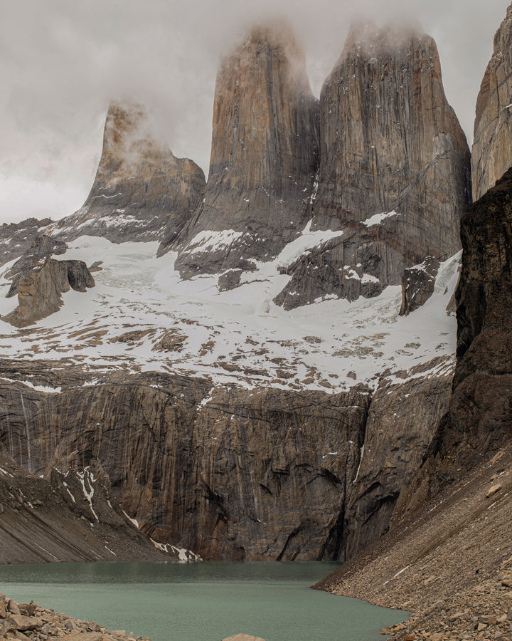 Majestic, towering rock formations rise from a snow-covered landscape, partially shrouded by misty clouds, with a turquoise glacial lake in the foreground.