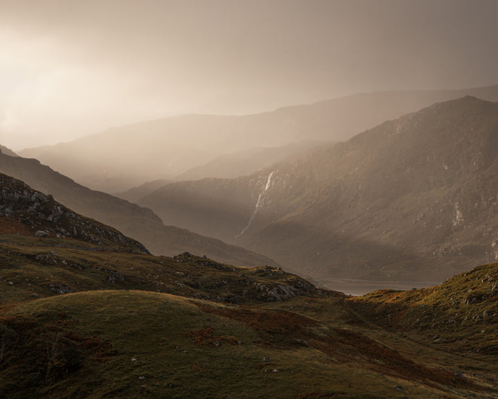 Rolling, grassy hills and distant mountains bathed in gentle, diffused sunlight, creating a moody and atmospheric landscape.