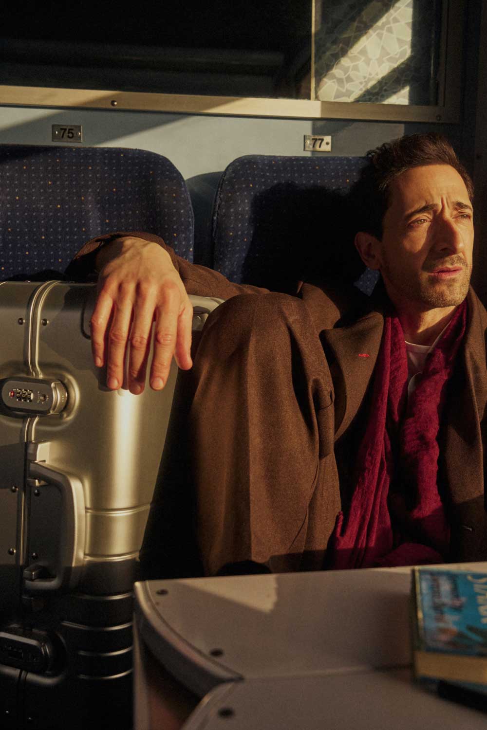 Adrien Brody sits by a train window, bathed in warm sunlight. He wears a brown coat and a deep red scarf, gazing outside with a contemplative expression. His hand rests on a Monos Aluminum Carry-On Plus suitcase in Aspen Silver beside him. The train’s blue seats and numbered labels 