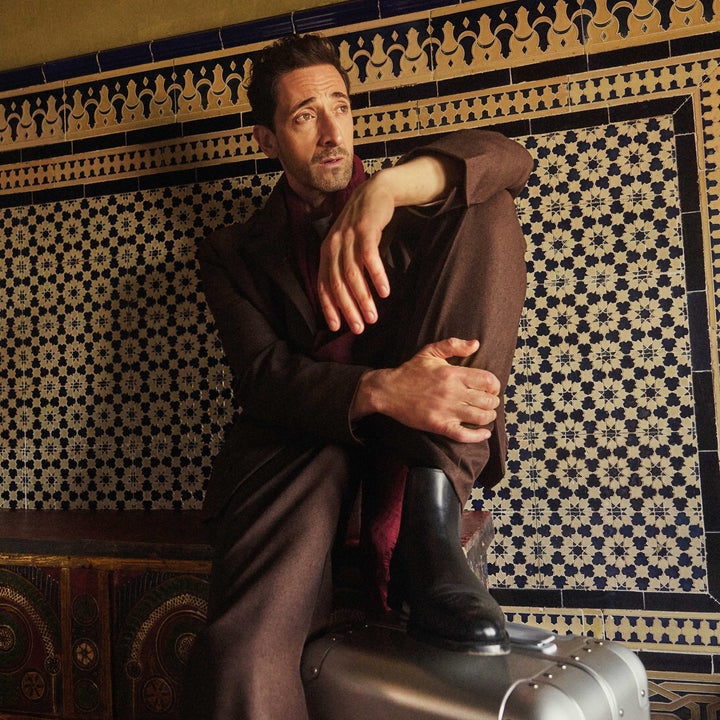 Adrien Brody wearing a brown coat and red scarf sits against an intricately tiled wall with geometric patterns. His boot rests on a Monos aluminum suitcase in aspen silver as he gazes into the distance with a contemplative expression.