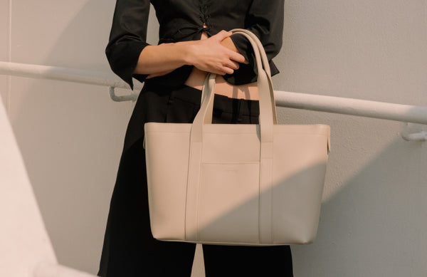 A person holding a metro tote in ivory (vegan leather), standing against a minimalist white background.