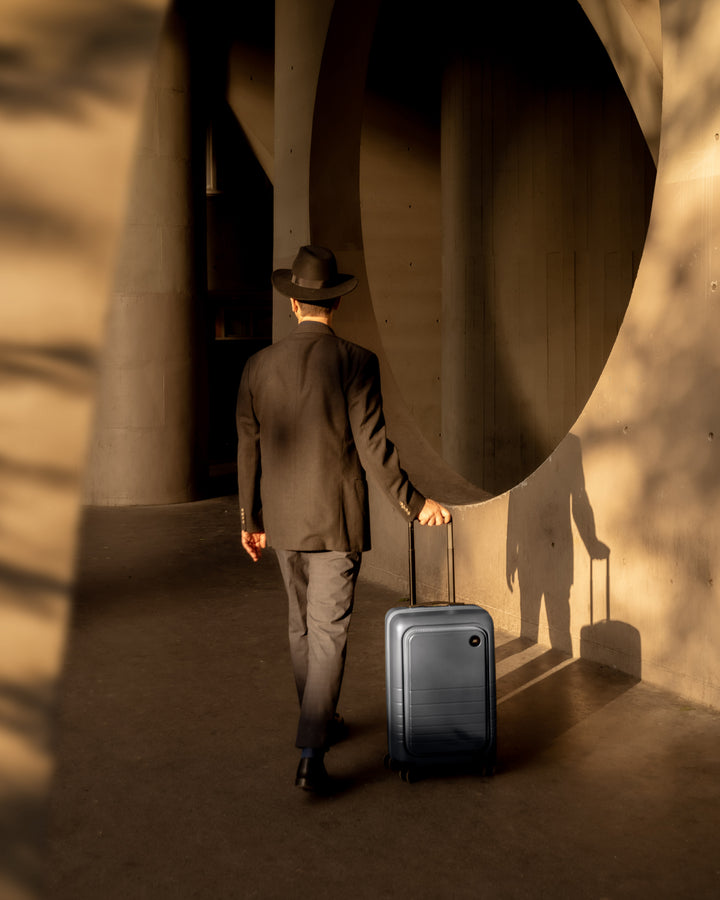 A man dressed in dark clothing strolls through a concrete structure, pulling a carry-on pro in ocean blue, with warm sunlight casting dramatic shadows.