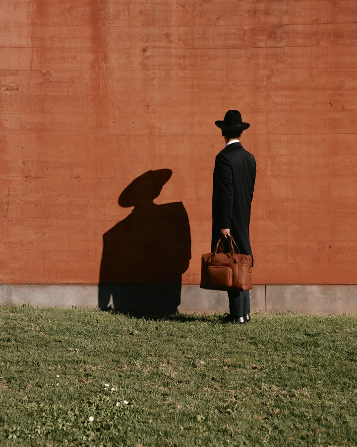 A man in a black outfit and hat stands facing a red wall, casting a distinct shadow, with a metro duffel in mahogany  (vegan leather) in hand, creating a stylish, minimalist scene.