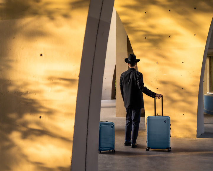 A man stands with a carry-on pro and check-in medium in ocean blue in a striking architectural structure, with golden light illuminating the background, creating a warm, modern atmosphere.