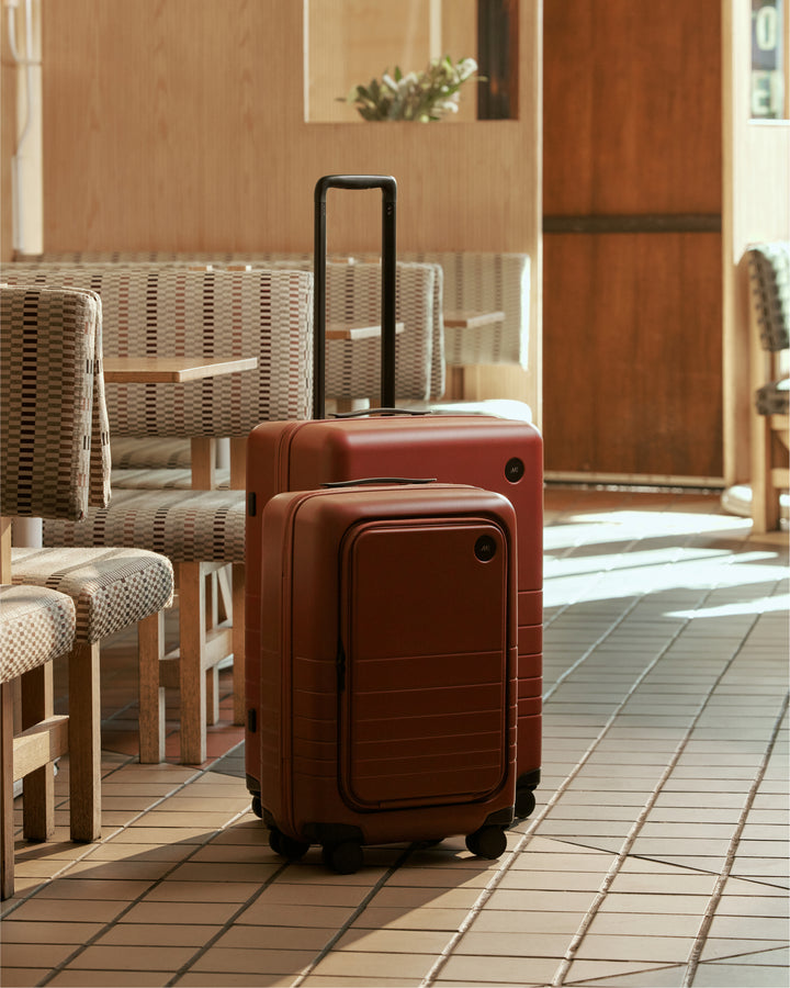 A carry-on pro and check-in medium in terracotta stand in a warm, inviting café setting with tiled floors and woven-patterned seating.