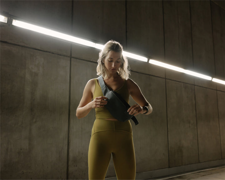 A woman in a workout outfit stands in an industrial hallway with concrete walls and bright overhead lighting, adjusting a metro sling in dover grey on her chest.