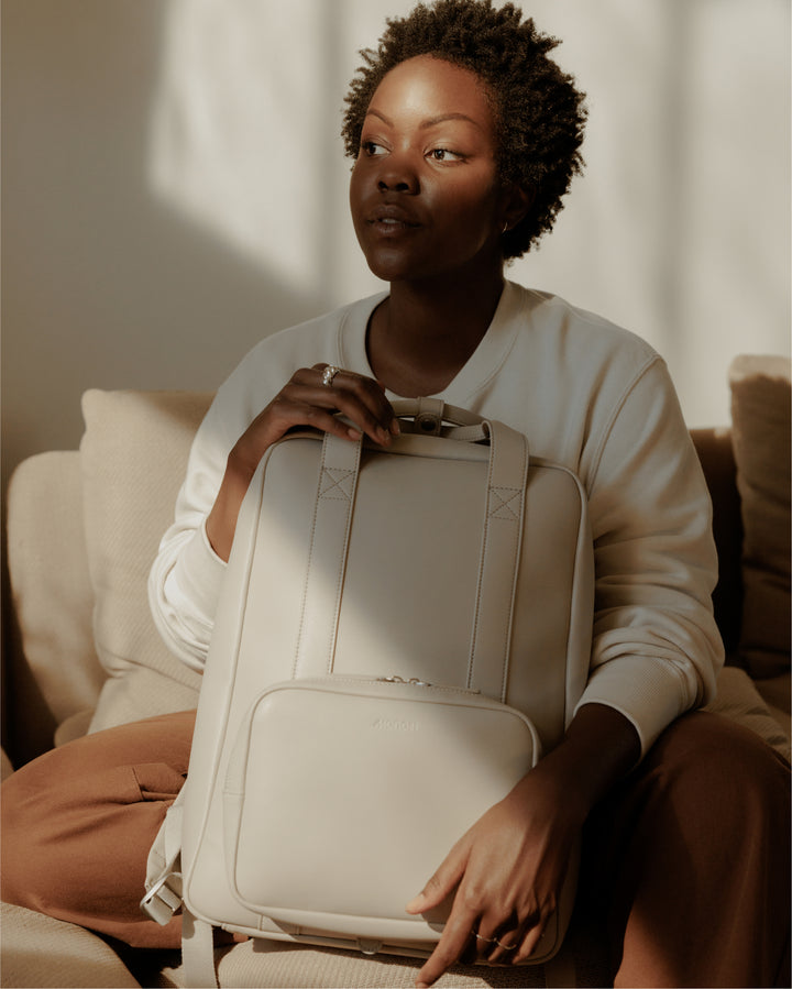 A person sitting on a cozy couch, holding a metro backpack in ivory (vegan leather) with a soft, contemplative expression as natural light casts gentle shadows across their face.