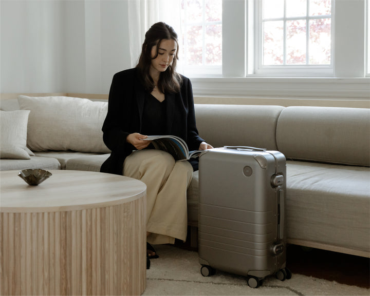 A woman in a sophisticated, minimalistic living room, reading a magazine with hybrid carry-on in silver by her side, surrounded by soft, neutral-toned furnishings.