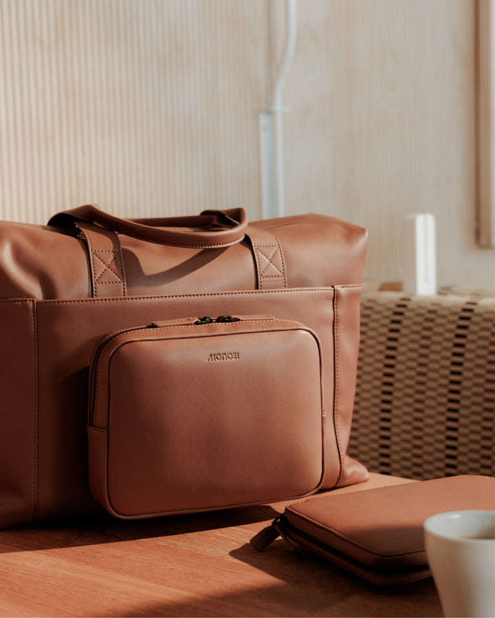 A close-up of a metro duffel in mahogany (vegan leather) with a matching smaller pouch, placed on a wooden table in a bright, sunlit room.