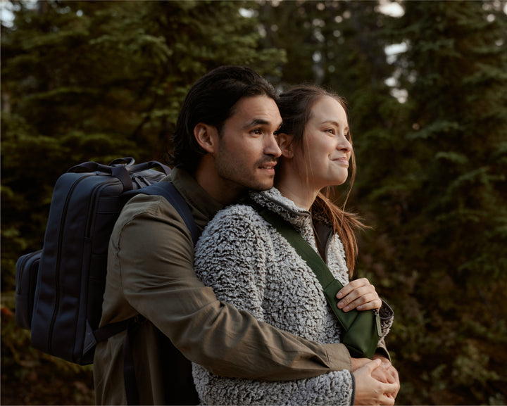 A couple standing in a forest setting, embracing warmly as they look into the distance with serene expressions. The man carries a metro backpack in oxford blue, and the woman with a metro sling in juniper green holds onto his arm, both dressed in cozy outdoor wear.