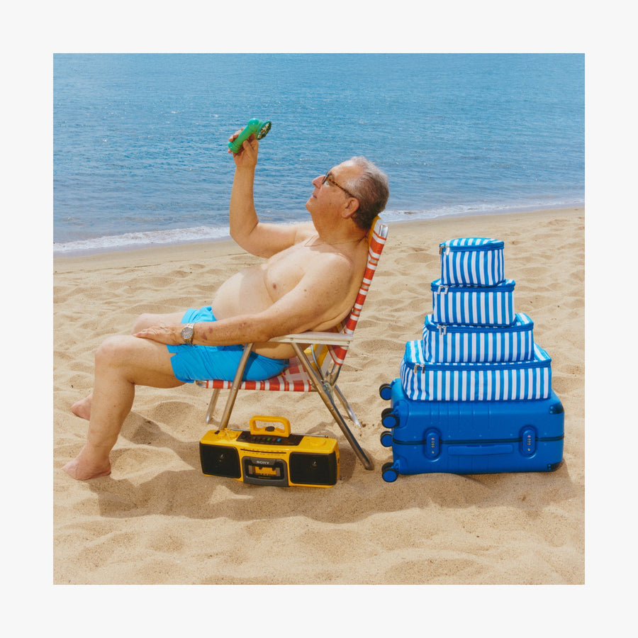 Coastal Blue | Man relaxing on a chair, cooling off with a portable fan beside a stack of Coastal Blue Hybrid suitcases and Poplin Stripe Coastal Blue Packing Cubes in various sizes