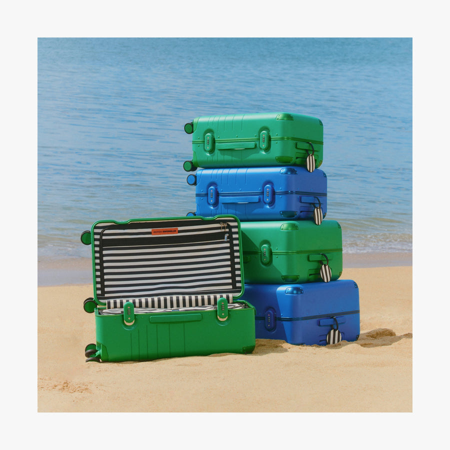 Jungle Green | Stack of Jungle Green and Coastal Blue Hybrid suitcases on the sand, with one open suitcase revealing its striped interior