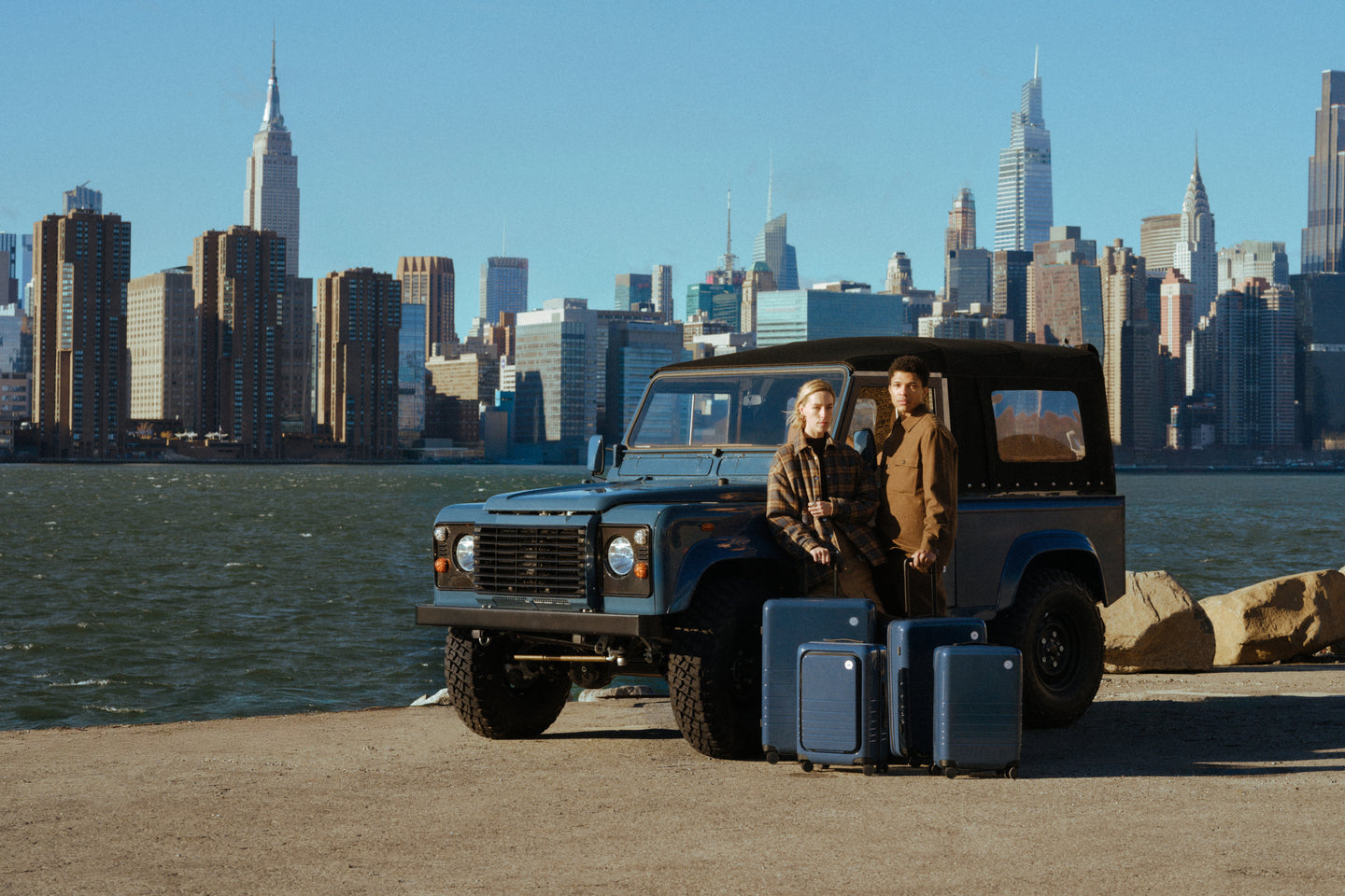 Two people standing next to a vintage SUV with suitcases in front of a city skyline.