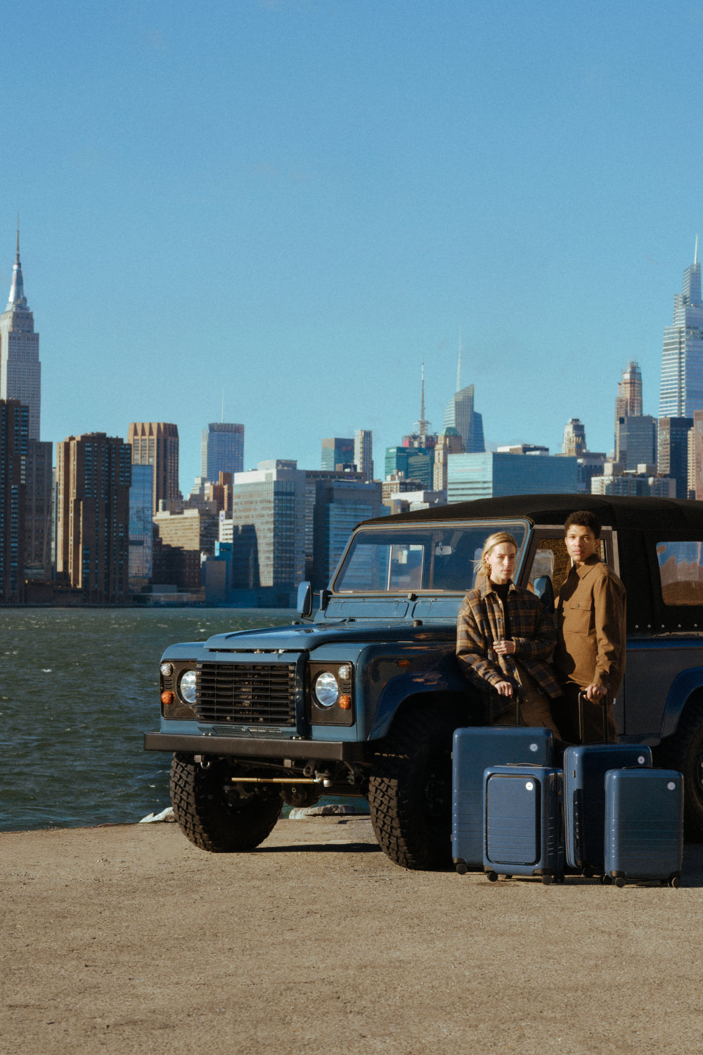 Two people with suitcases standing next to a Land Rover Defender by a waterfront with a city skyline in the background.