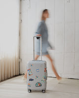 The Monos Hybrid Carry-On in silver, decorated with Destination Stickers, rolls across a sunlit floor beside a blurred figure in blue.