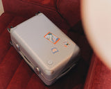 The Monos Hybrid Carry-On in silver rests on a deep red sofa, its shell decorated with Destination Stickers that hint at journeys ahead.