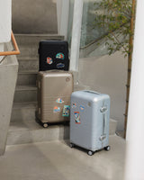 Three Monos Hybrid Carry-On suitcases in Obsidian, Champagne, and Silver stand on concrete steps, each personalized with Destination Stickers.