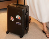 The Monos Hybrid Carry-On in Obsidian, accented with Destination Stickers, stands beside a traveler in a flowing white dress and strappy sandals.