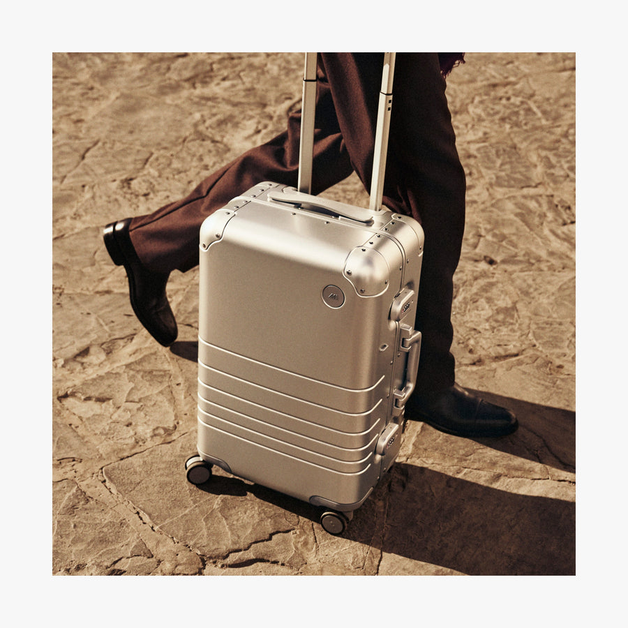 Champagne Gold | A close-up of a Monos aluminum carry-on plus suitcase in aspen silver. A person wearing dark brown trousers and polished black shoes walks alongside it on a textured stone pavement, with warm sunlight casting soft shadows.