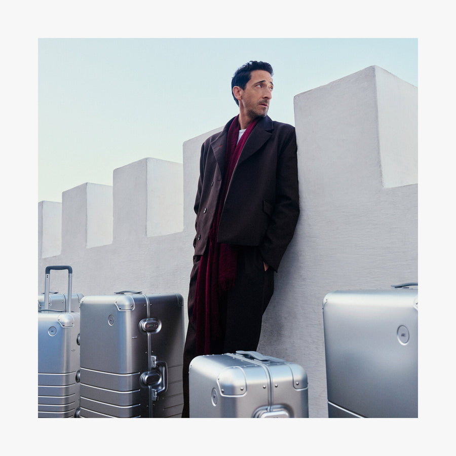 Caviar Black | Adrien Brody leans against a white fortress-like wall, gazing into the distance, with multiple Monos aluminum suitcases in aspen silver around him.