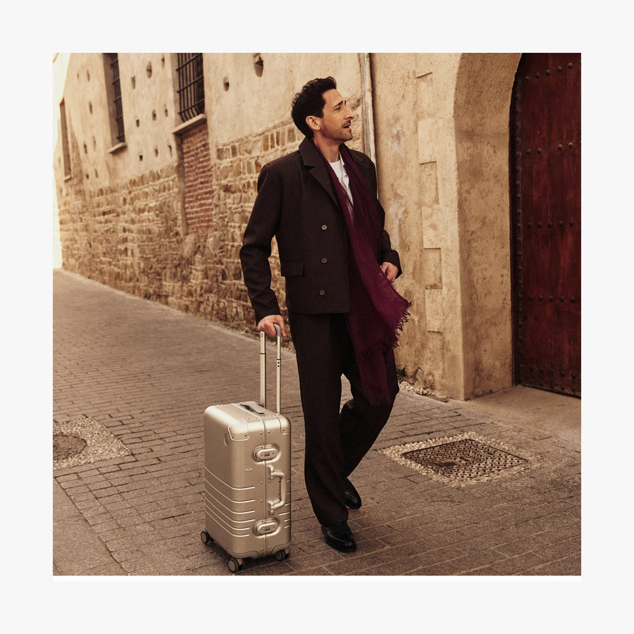 Aspen Silver | Adrien Brody dressed in a dark brown suit, white shirt, and a deep burgundy scarf walks down a historic cobblestone street while pulling a Monos Aluminum carry-on plus suitcase in aspen silver. The background features an old stone building with a wooden door, adding to the image's timeless, travel-inspired aesthetic.