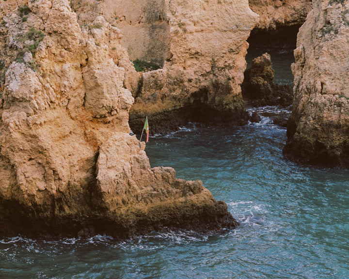 A rugged coastal cliff with clear blue-green waves crashing against it, highlighting the raw beauty of nature with a small colourful flag adding a hint of human presence.