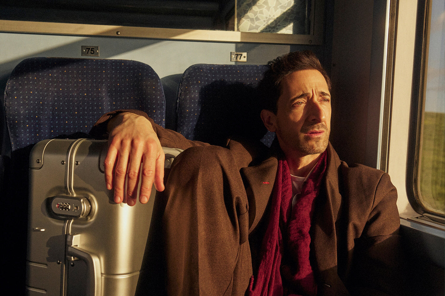 Adrien Brody sits by a train window, bathed in warm sunlight. He wears a brown coat and a deep red scarf, gazing outside with a contemplative expression. His hand rests on a Monos Aluminum Carry-On Plus suitcase in Aspen Silver beside him. The train’s blue seats and numbered labels 