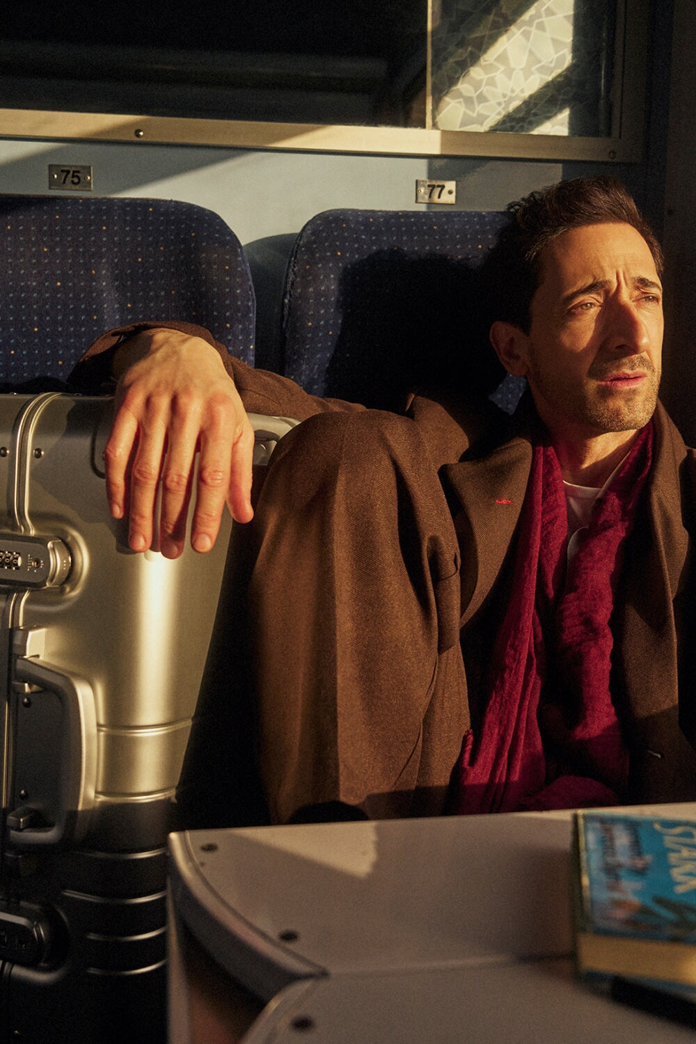 Adrien Brody sits by a train window, bathed in warm sunlight. He wears a brown coat and a deep red scarf, gazing outside with a contemplative expression. His hand rests on a Monos Aluminum Carry-On Plus suitcase in Aspen Silver beside him. The train’s blue seats and numbered labels 