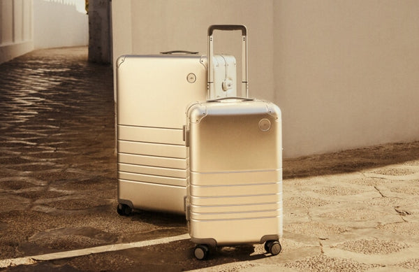 Two Monos aluminum suitcases in Aspen Silver, one in Check-In Medium and one in Carry-On Plus, stand on a textured stone path beside a white-walled building. The sunlight casts a warm glow, highlighting their sleek metallic finish.