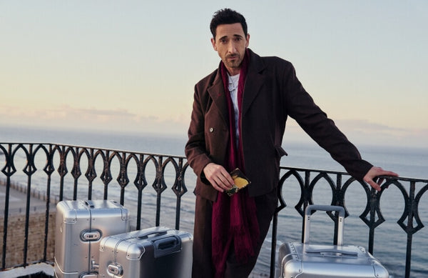 Adrien Brody with short dark hair and a red scarf leans against a wrought iron railing overlooking the ocean at dusk. He wears a brown coat and holds sunglasses in one hand, with several Monos aluminum suitcases in Aspen Silver beside him.