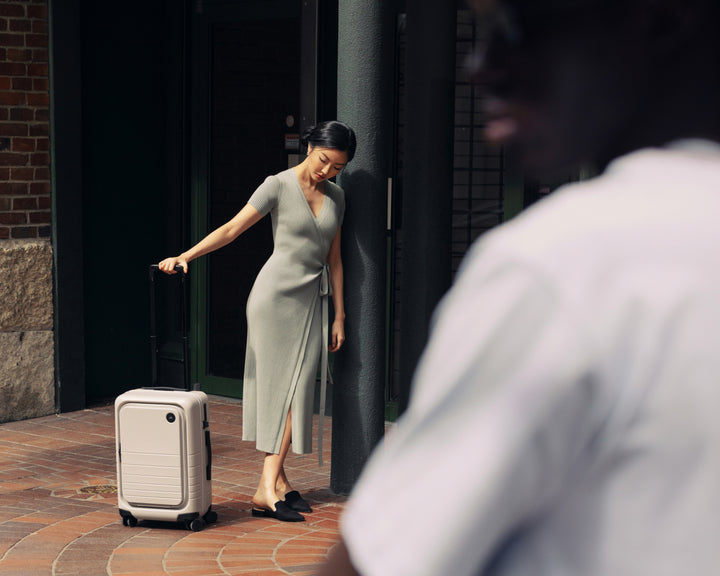 A woman leans against a pole, looking down thoughtfully to her Carry-On Pro in Desert Taupe while the foreground features a man in focus wearing a white shirt.