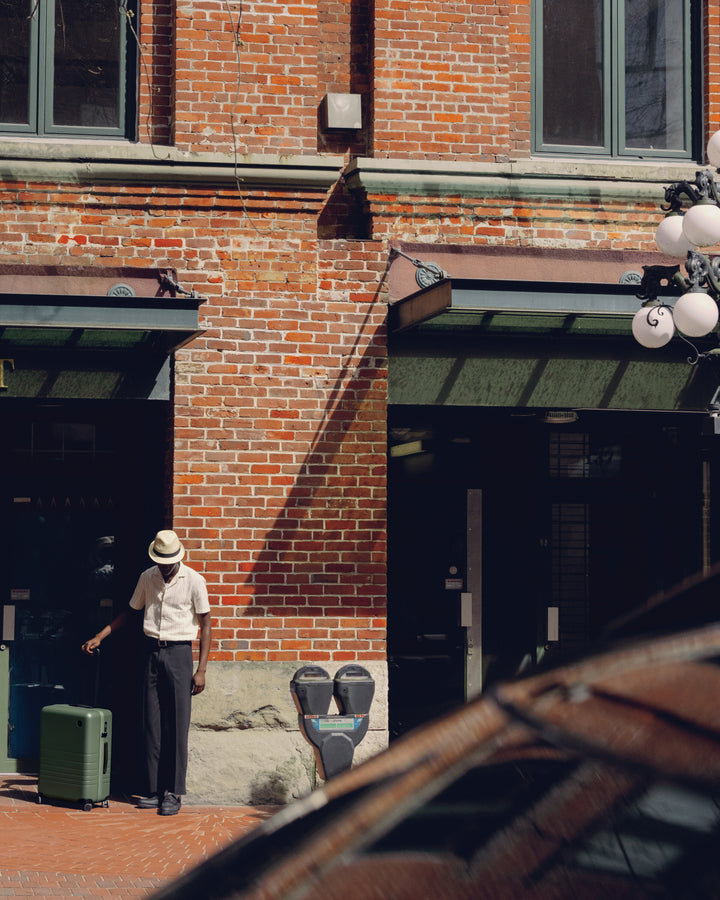 A man stands outside a brick building with a Check-In Medium in Olive Green beside him, casting a shadow in the sunlight.