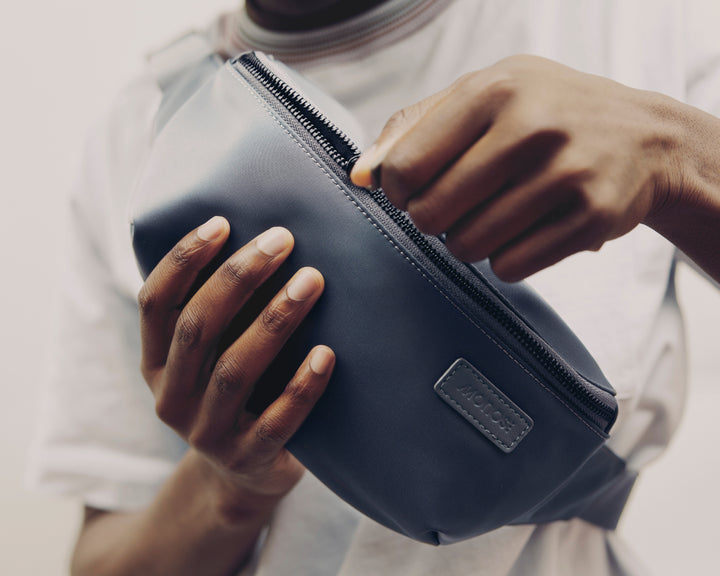 A close-up of a person’s hands unzipping a Metro Sling in Oxford Blue, highlighting the sleek design and logo of the bag.