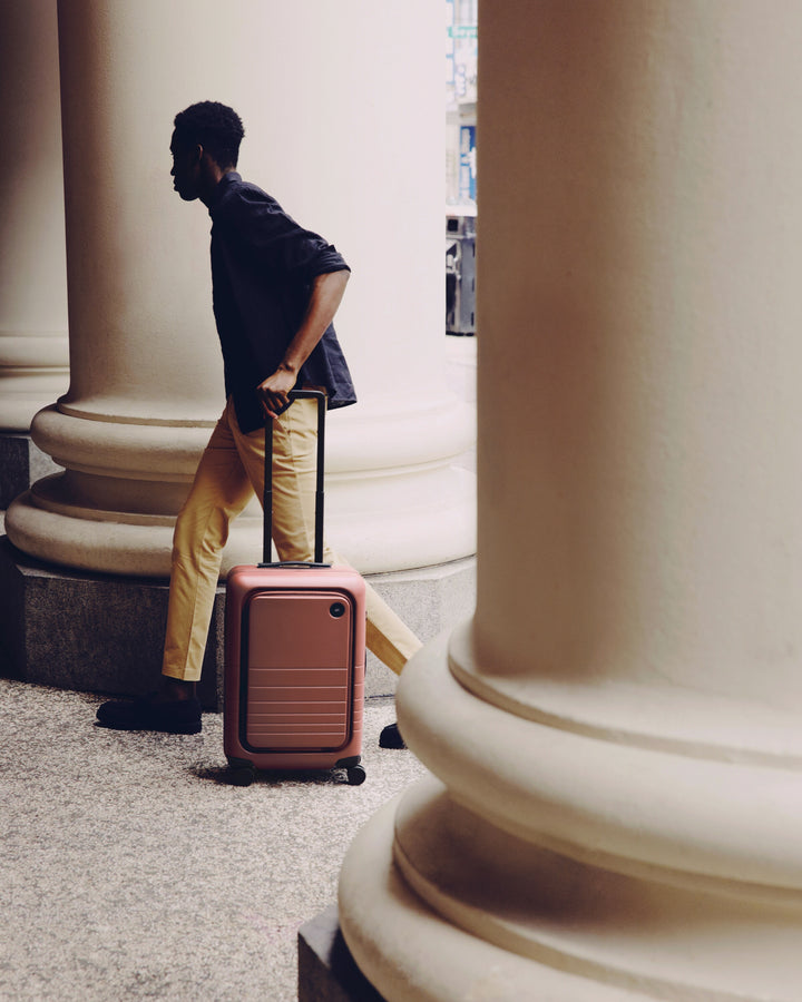 A man walks past large columns in a classical building, pulling a Carry-On Pro in Terracotta beside him.