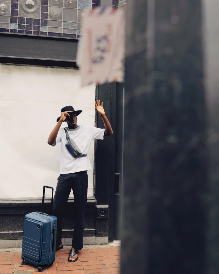 A man poses playfully outside with a Carry-On Pro in Ocean Blue, adjusting his hat, while a nearby sign is slightly out of focus in the foreground.