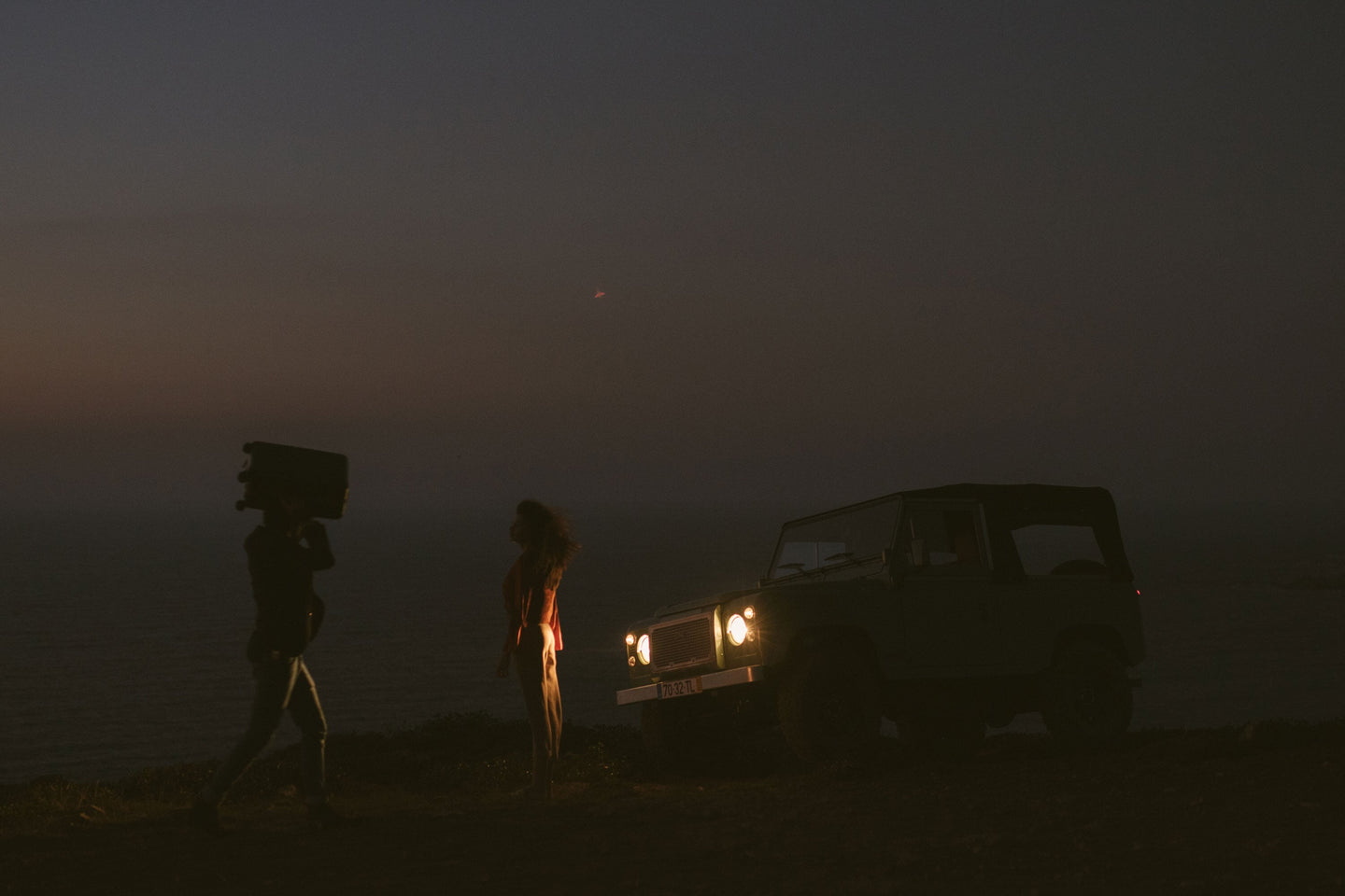 A dimly lit scene at dusk featuring a vintage SUV with its headlights on, parked near the edge of a cliff overlooking the ocean. In the foreground, a man carries a carry-on luggage on his shoulder, walking toward the vehicle, while a woman stands facing the sea, her silhouette softly illuminated by the headlights.