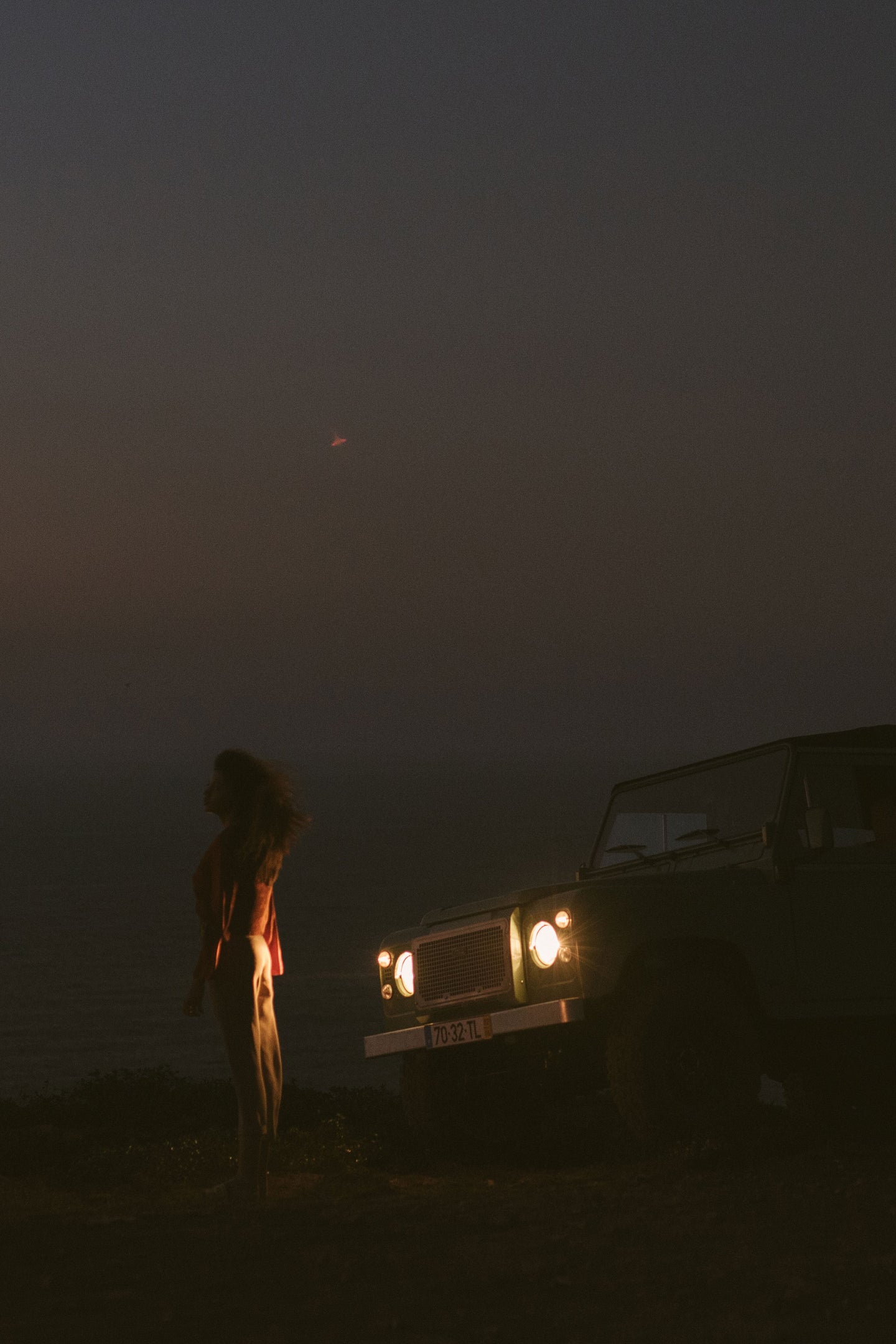 A dimly lit scene at dusk featuring a vintage SUV with its headlights on, parked near the edge of a cliff overlooking the ocean. A woman stands facing the sea, her silhouette softly illuminated by the headlights.