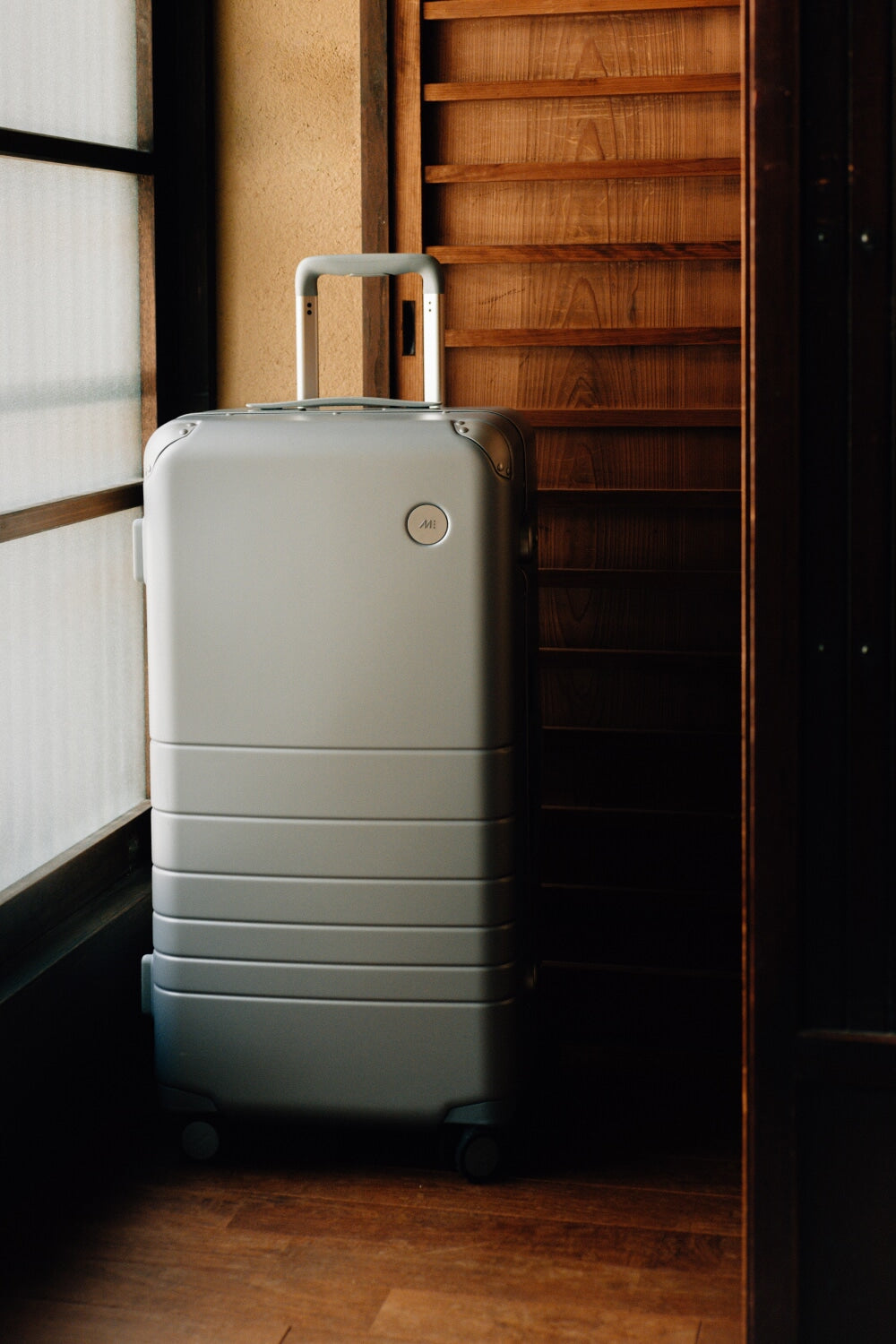 A close-up of a hybrid trunk in silver standing against a traditional wooden backdrop with soft natural light streaming through a nearby window. The suitcase's smooth, modern design contrasts with the rustic wooden slats behind it.