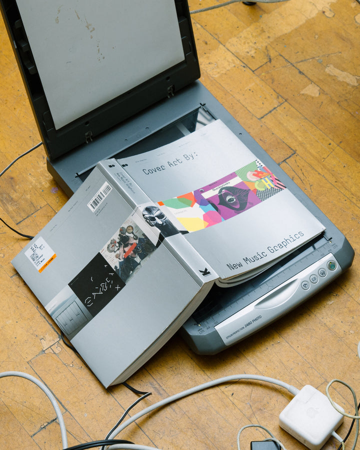 A flatbed scanner with an open book of music graphics, showcasing colourful album art. The setup suggests an exploration of album cover design, capturing the intersection of music and visual art.