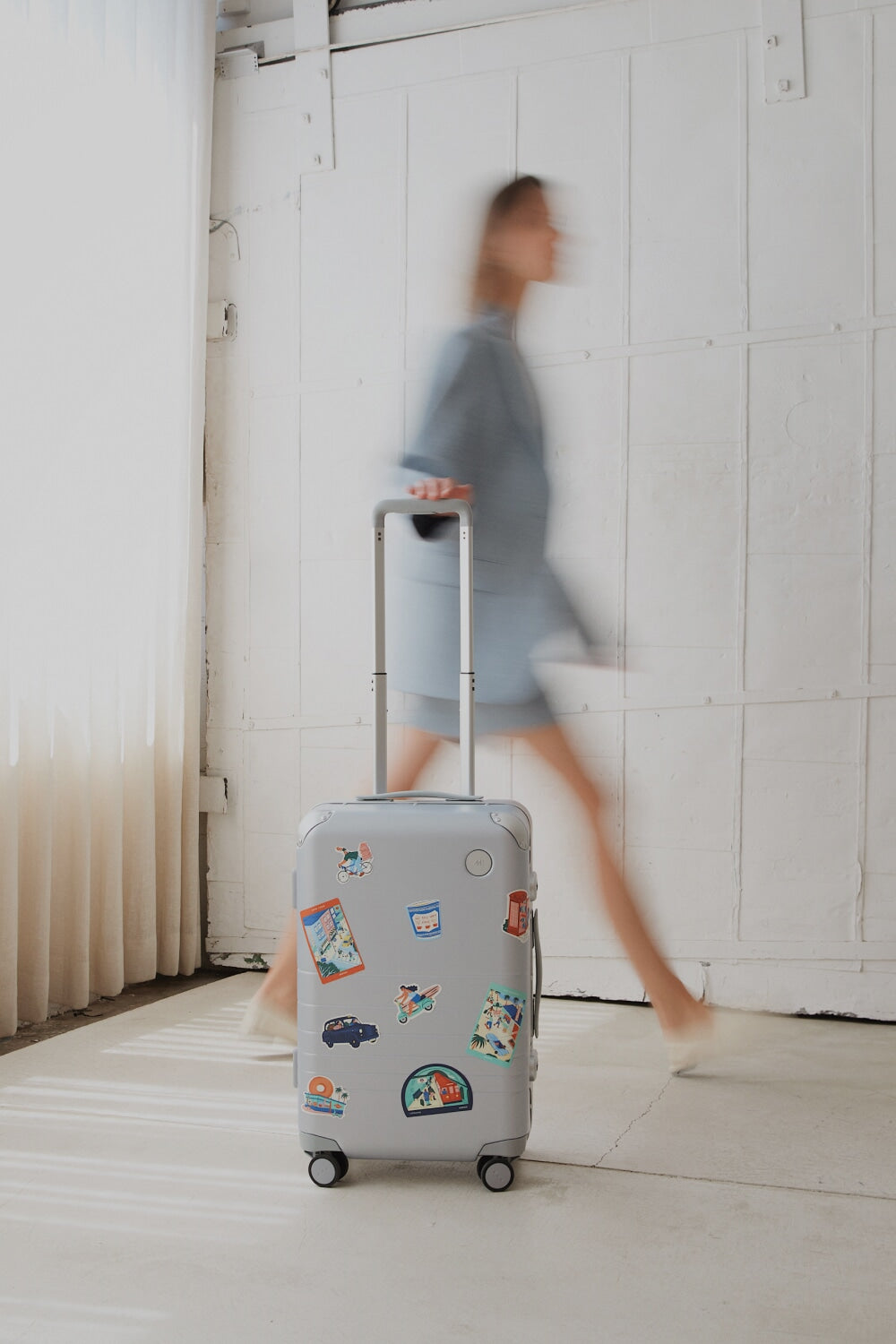 The Monos Hybrid Carry-On in silver, decorated with Destination Stickers, rolls across a sunlit floor beside a blurred figure in blue.