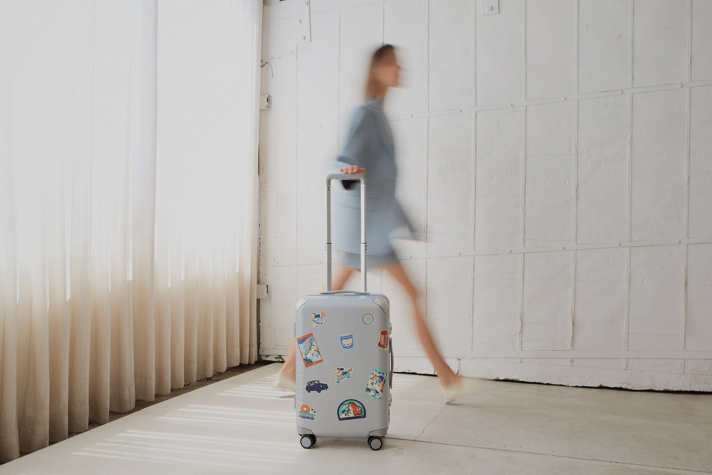The Monos Hybrid Carry-On in silver, decorated with Destination Stickers, rolls across a sunlit floor beside a blurred figure in blue.