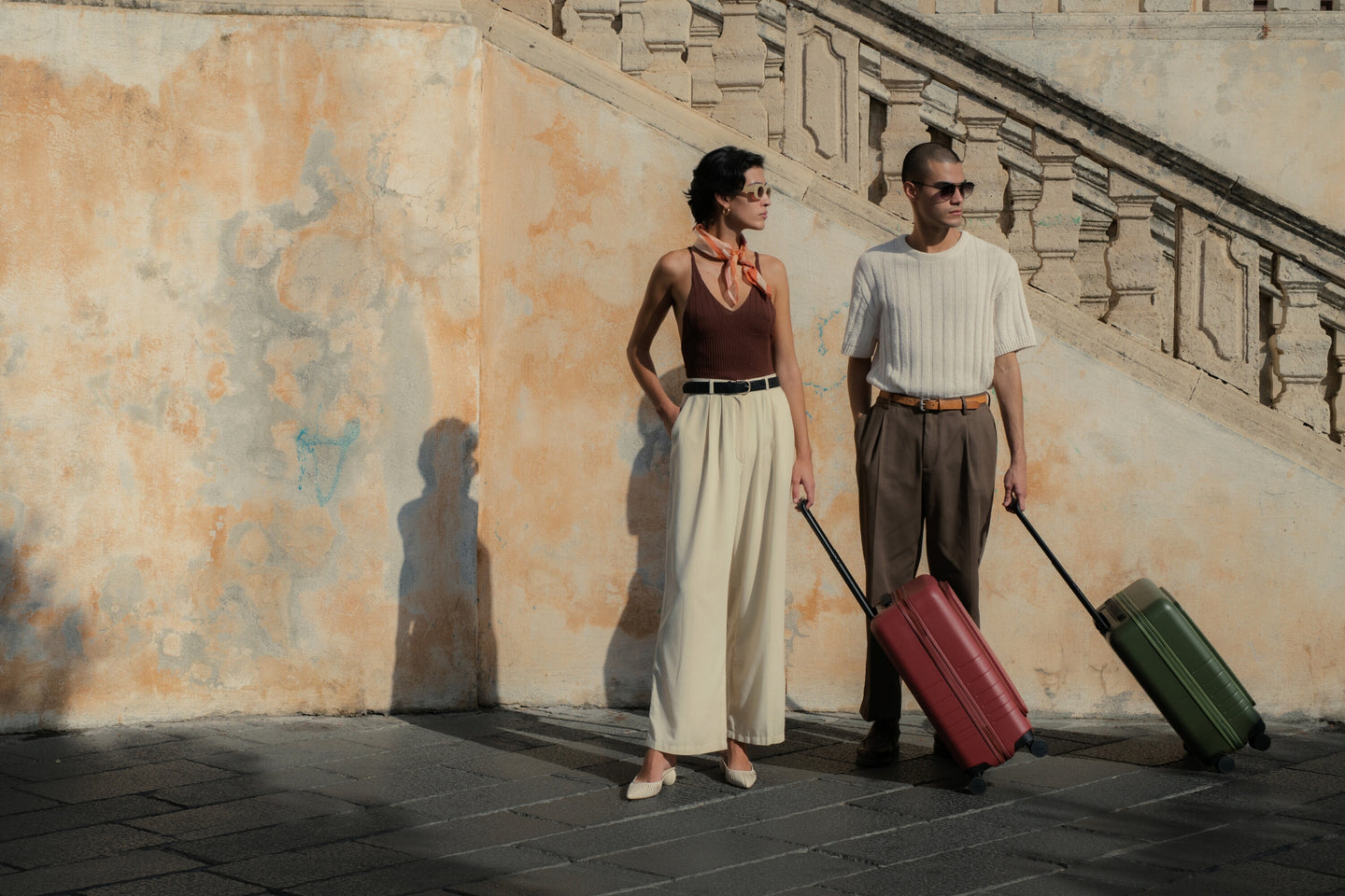 A couple stands side by side under golden hour light, each with a Monos carry-on in hand—sleek, intentional travel against a backdrop of timeless architecture.