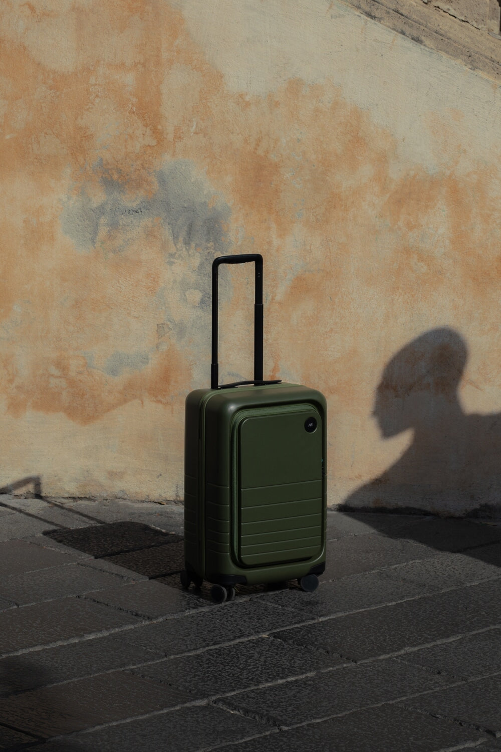 A solo Monos carry-on Pro in olive green rests against a sunlit stucco wall—shadows, texture, and stillness meet purposeful design.