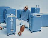 A model in a blue sweater sits among various sizes of Hybrid luggage in Elemental Blue on a light blue background.