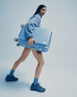 A model in a blue fleece jacket and shorts strides forward carrying a Hybrid Carry-On in Elemental Blue against a light blue backdrop.